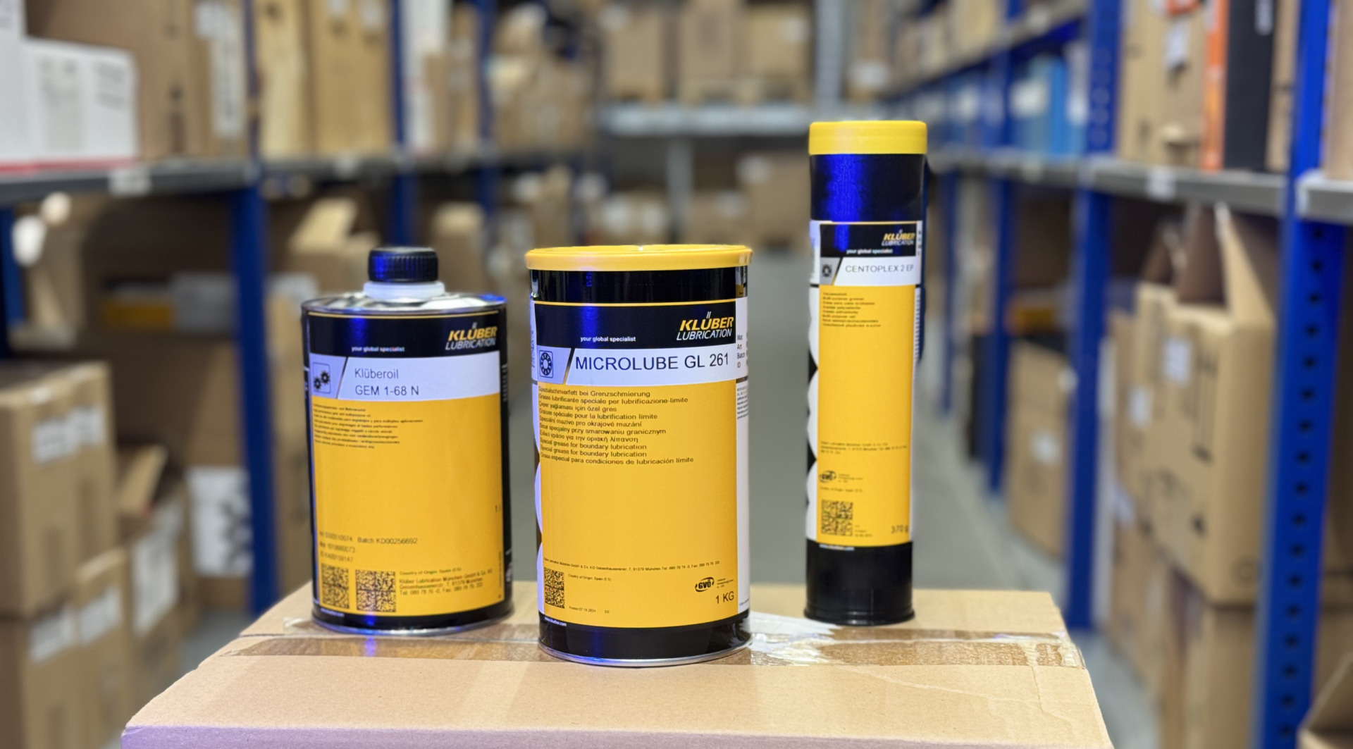 Drei Schmierstoffprodukte von Klüber Lubrication stehen auf einem Karton in einem Lagerregal.