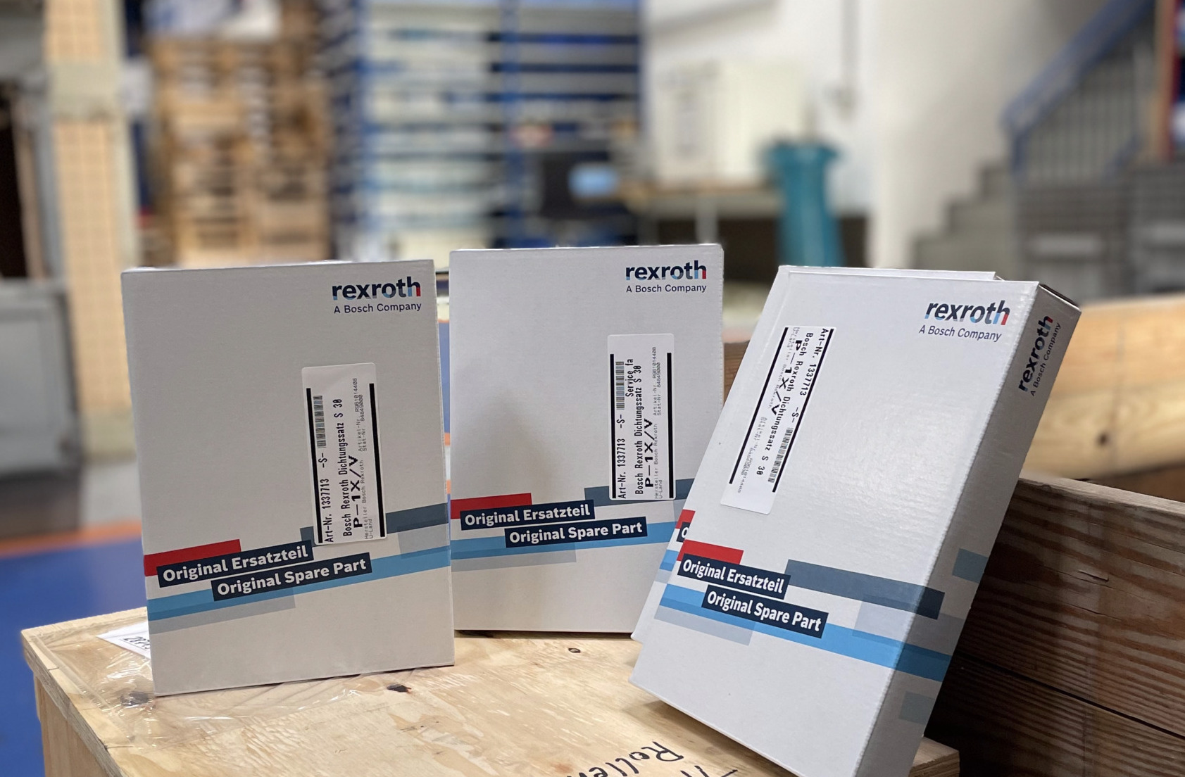 Drei Original-Ersatzteilverpackungen von Bosch Rexroth stehen auf einer Holzkiste in einem Lagerraum.
