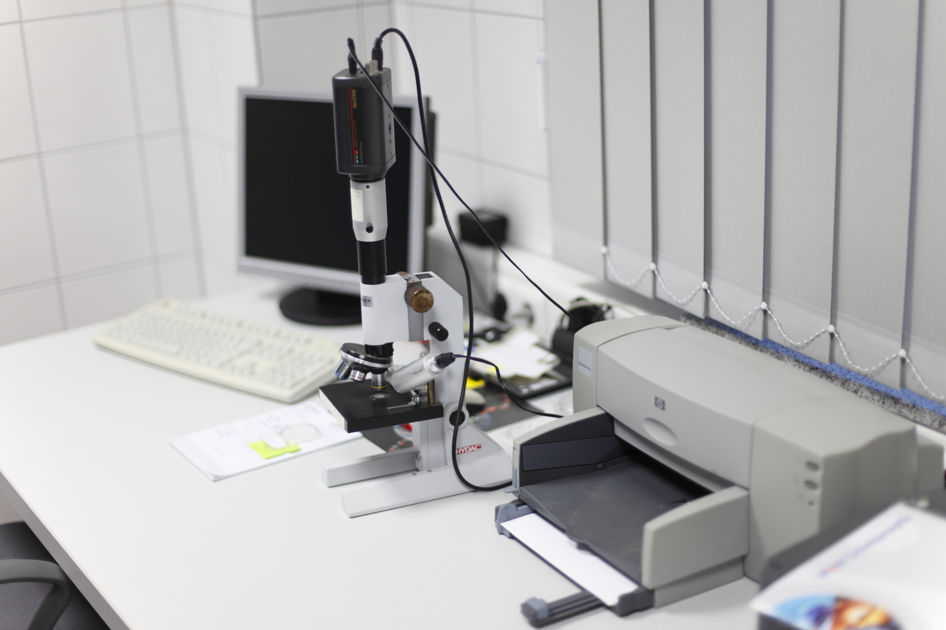 Ein digital ausgestatteter Prüfplatz mit Mikroskop, Kameraeinheit, Computer und Drucker in einem Laborraum.